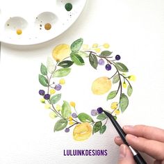 someone is painting a watercolor wreath with yellow and purple flowers on white paper next to a paintbrush