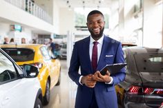 a man in a suit and tie standing next to some cars at a dealership