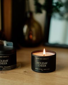 two candles sitting on top of a wooden table next to a bottle of holiday cheer