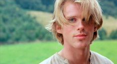 a young man with blonde hair and blue eyes looks at the camera while standing in front of a green field