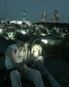 a man and woman sitting on top of a roof looking at a cell phone in the dark