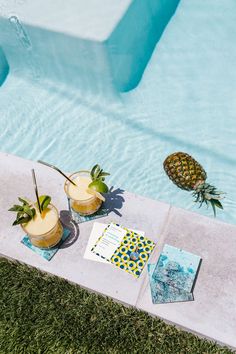 two drinks sitting on the edge of a swimming pool next to a pineapple drink