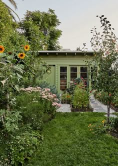 a small green building surrounded by trees and flowers in the yard with sunflowers