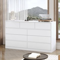 a white dresser sitting in front of a window next to a table with a vase on it