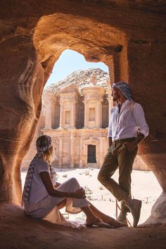 a man and woman sitting on the ground in front of a rock formation with a building in the background