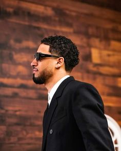 a man in a suit and sunglasses looking off into the distance with a wooden wall behind him