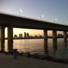 a bridge that is over some water with lights above it and buildings in the background