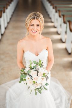 a woman in a wedding dress holding a bouquet and smiling at the camera while standing in front of rows of pews