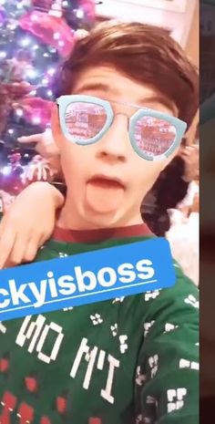 a young boy wearing sunglasses with the words skyisboss on it in front of a christmas tree