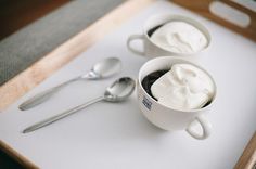 two mugs filled with whipped cream sit on a cutting board next to spoons