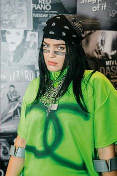 a woman with black hair wearing a green shirt and bandana, standing in front of a poster