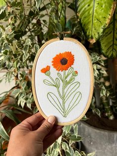 a person holding up an embroidered orange flower in front of a potted plant with green leaves