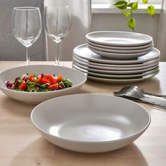 a table topped with white plates and bowls filled with salad next to wine glasses on top of a wooden table