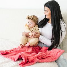 a woman holding a baby sitting on top of a bed next to a pink blanket