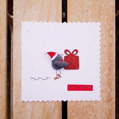 a card with a bird holding a red gift box on it's back, sitting on top of a wooden bench