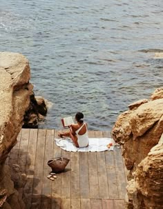 a woman sitting on a dock reading a book next to the ocean with ducks nearby