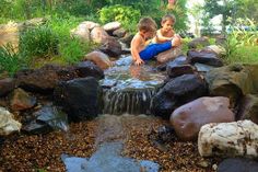 two young boys sitting on rocks playing in a pond with water flowing from it's sides