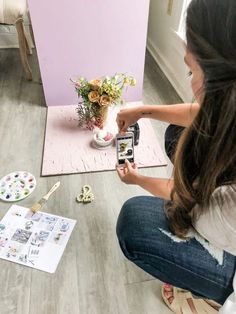 a woman sitting on the floor taking pictures with her cell phone and painting flowers in a vase