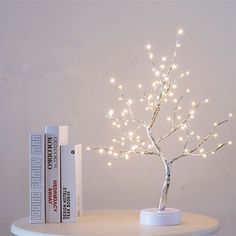 a small tree with white lights on top of a table next to books and a lamp