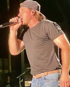 a man in grey shirt and jeans holding a microphone to his mouth while standing on stage