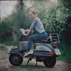 a woman riding on the back of a gray scooter down a dirt road