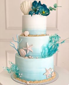 a three tiered cake decorated with sea shells and blue flowers on a white table