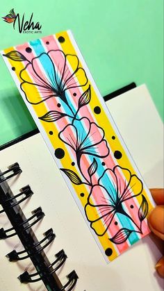 a hand holding a bookmark with flowers on it next to an open spiral notebook