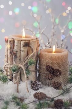 two candles are sitting in the snow next to some pine cones and evergreen branches with lights behind them