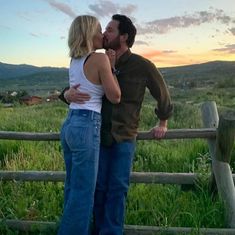 a man and woman kissing in front of a wooden fence with mountains in the background