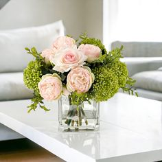 a glass vase filled with pink roses and greenery on a white table in a living room