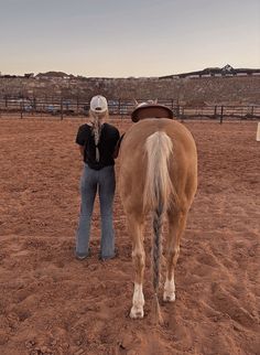 a woman standing next to a brown horse on top of a dirt field in front of a fence