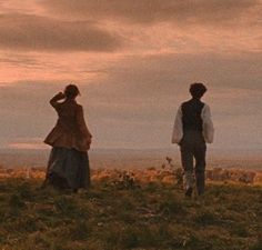 two people standing on top of a grass covered hill under a cloudy sky at sunset