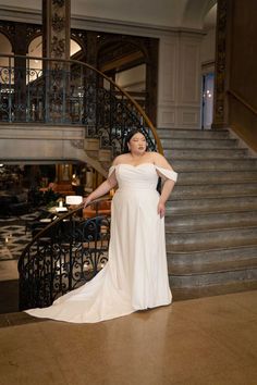 a woman standing in front of a staircase wearing a white wedding dress with off the shoulder sleeves