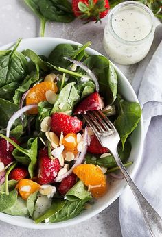 a salad with spinach, oranges and nuts in a white bowl next to a glass of dressing