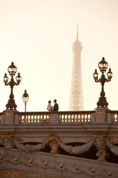 the eiffel tower is in the distance behind two people on a bridge with street lamps