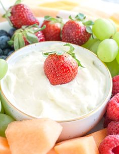 a white bowl filled with fruit and dip surrounded by grapes, melons, strawberries, and kiwis