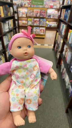 a hand holding a baby doll in a store aisle with bookshelves behind it