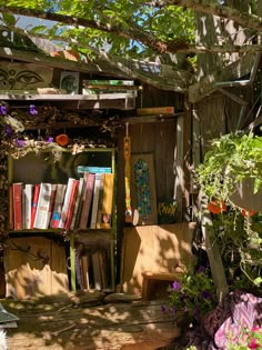 an old wooden book case filled with books under a tree in the shade, surrounded by flowers and plants