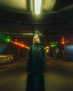 a woman standing in a parking garage wearing a long green coat and black pants with her head turned to the side