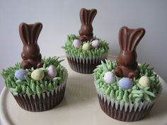 three cupcakes decorated with chocolate bunny ears and eggs in grass on a plate