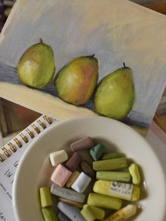 two pears and some crayons in a white bowl next to a drawing