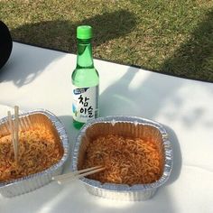 two containers of noodles with chopsticks next to a bottle of soda on a table