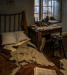 an unmade bed sitting in front of a window next to a desk with papers on it