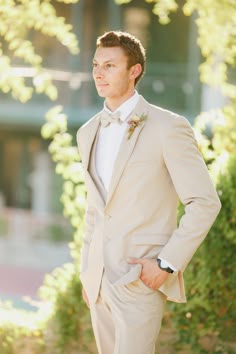 a man wearing a tan suit and bow tie standing in front of some bushes with his hands on his hips