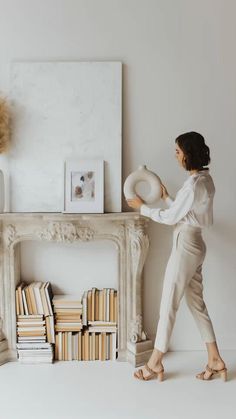 a woman standing in front of a white fireplace holding a hat and looking at books