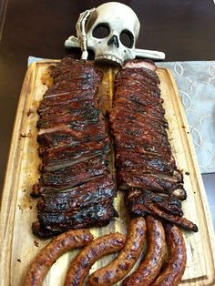 a wooden cutting board topped with ribs and sausages next to a skull on the table