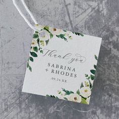 a white and green floral wedding thank you tag on a gray background with the words,'thank you sabrina + rhodes '