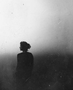 a black and white photo of a woman standing in the fog with her back to the camera