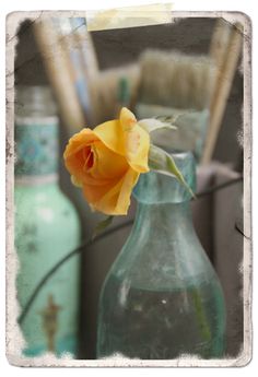 a yellow rose in a glass vase on a table next to other bottles and brushes