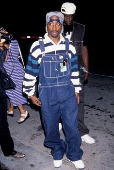 a man in overalls and a hat standing on the street with people behind him